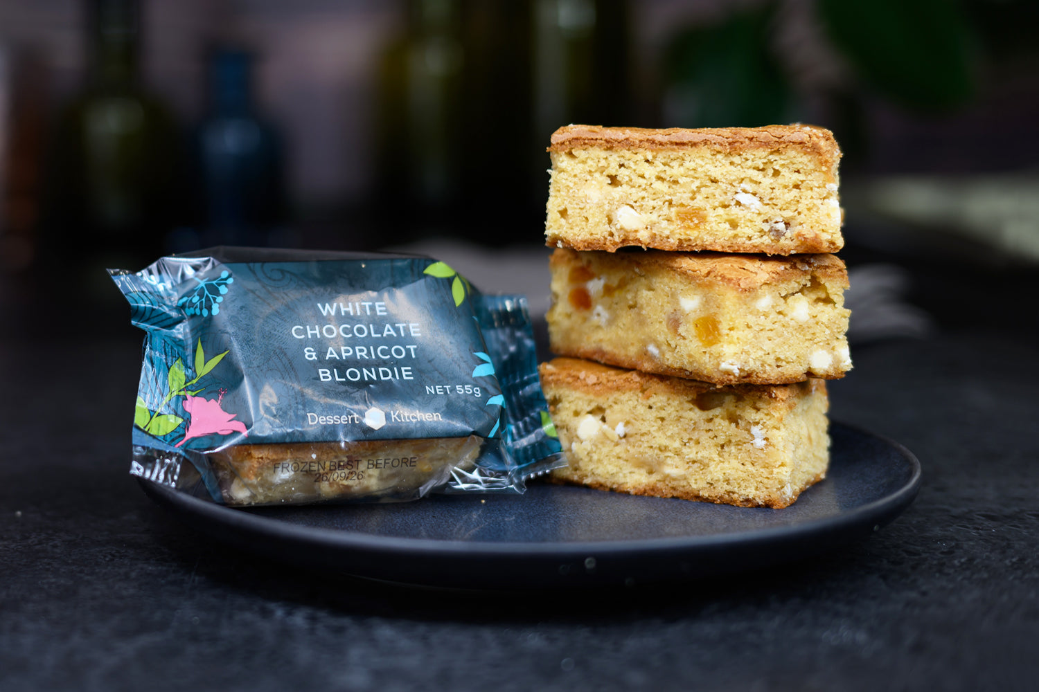Stack of white chocolate apricot blondies with a blue packaging on a dark surface Dessert Kitchen Partner Manufacturing