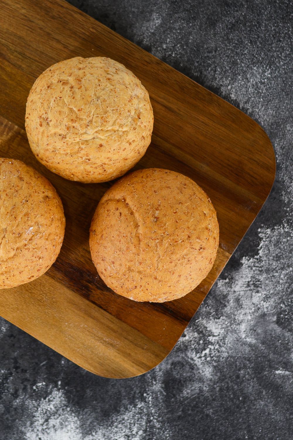 Three bread rolls or muffins on a wooden cutting board with a dark gray background. Dessert Kitchen Partner Manufacturing