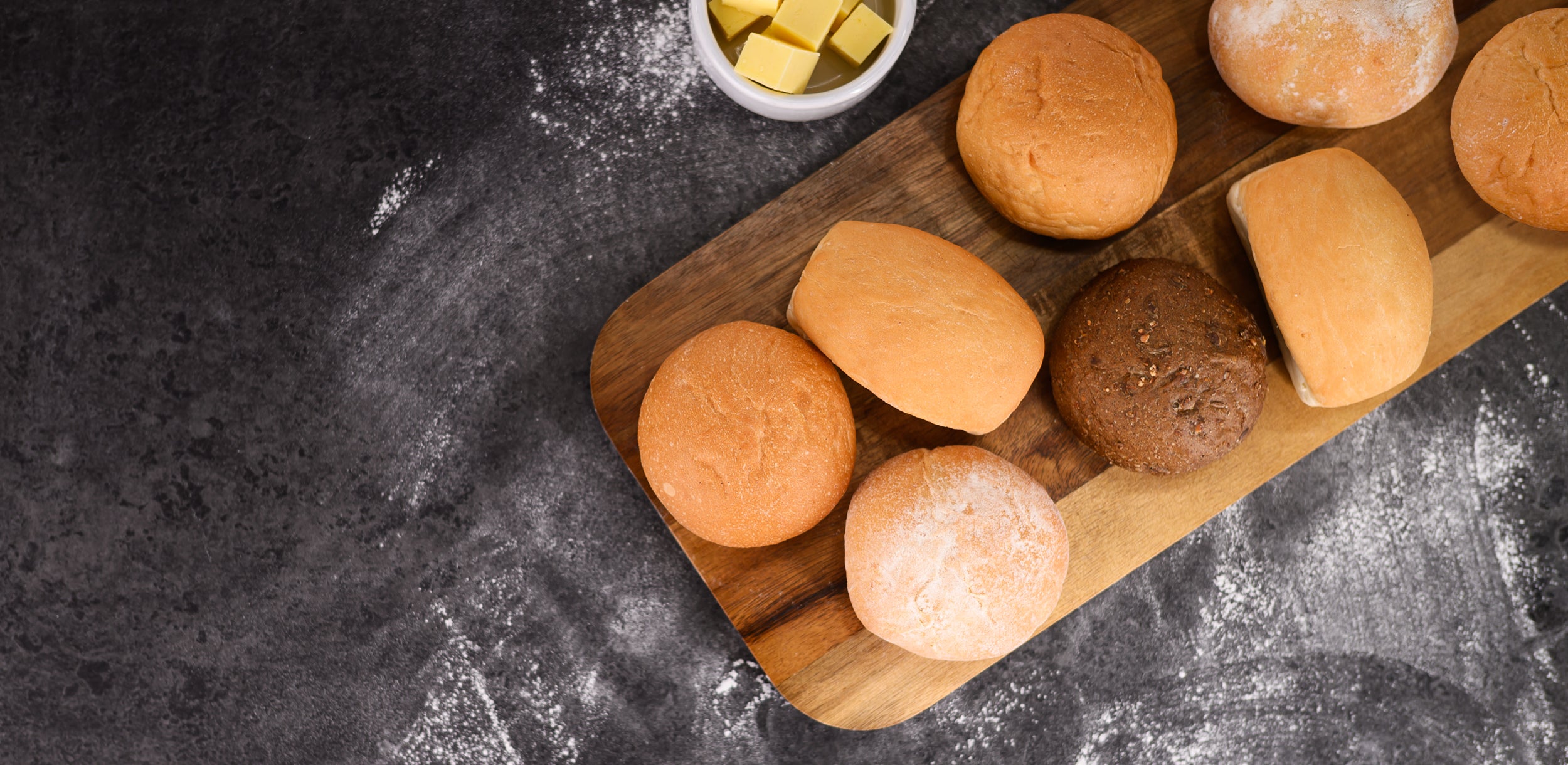 Assorted bread rolls on a wooden board with butter on a dark surface. Dessert Kitchen Breads & Rolls