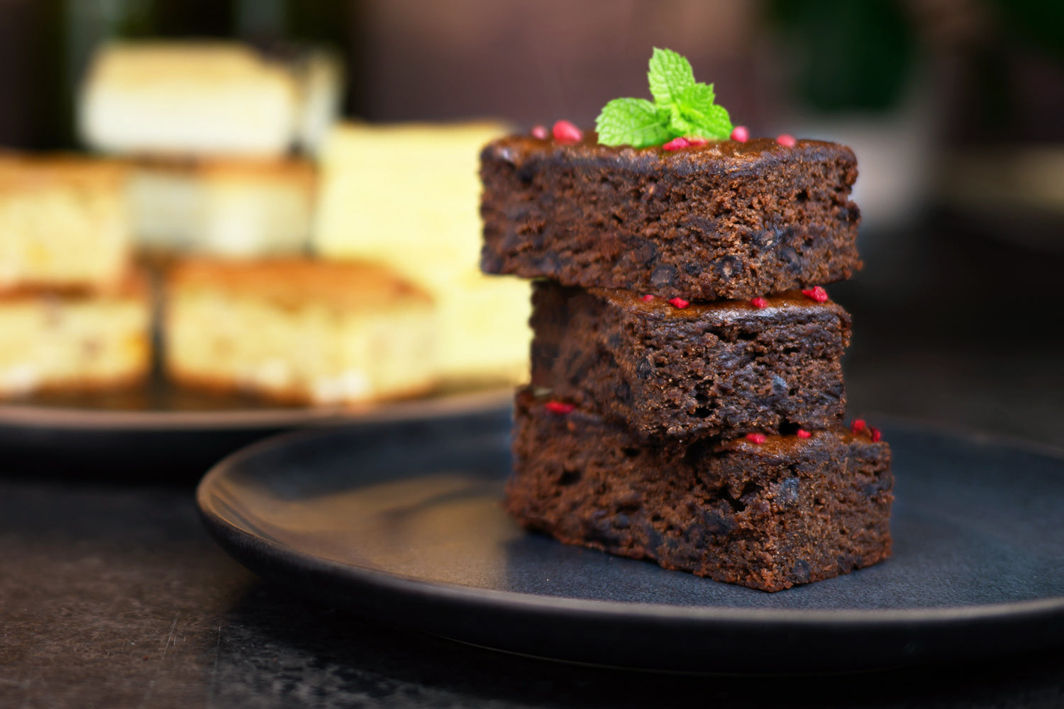 Stack of brownies on a black plate with a blurred dessert in the background. Dessert Kitchen Desserts & Brownies
