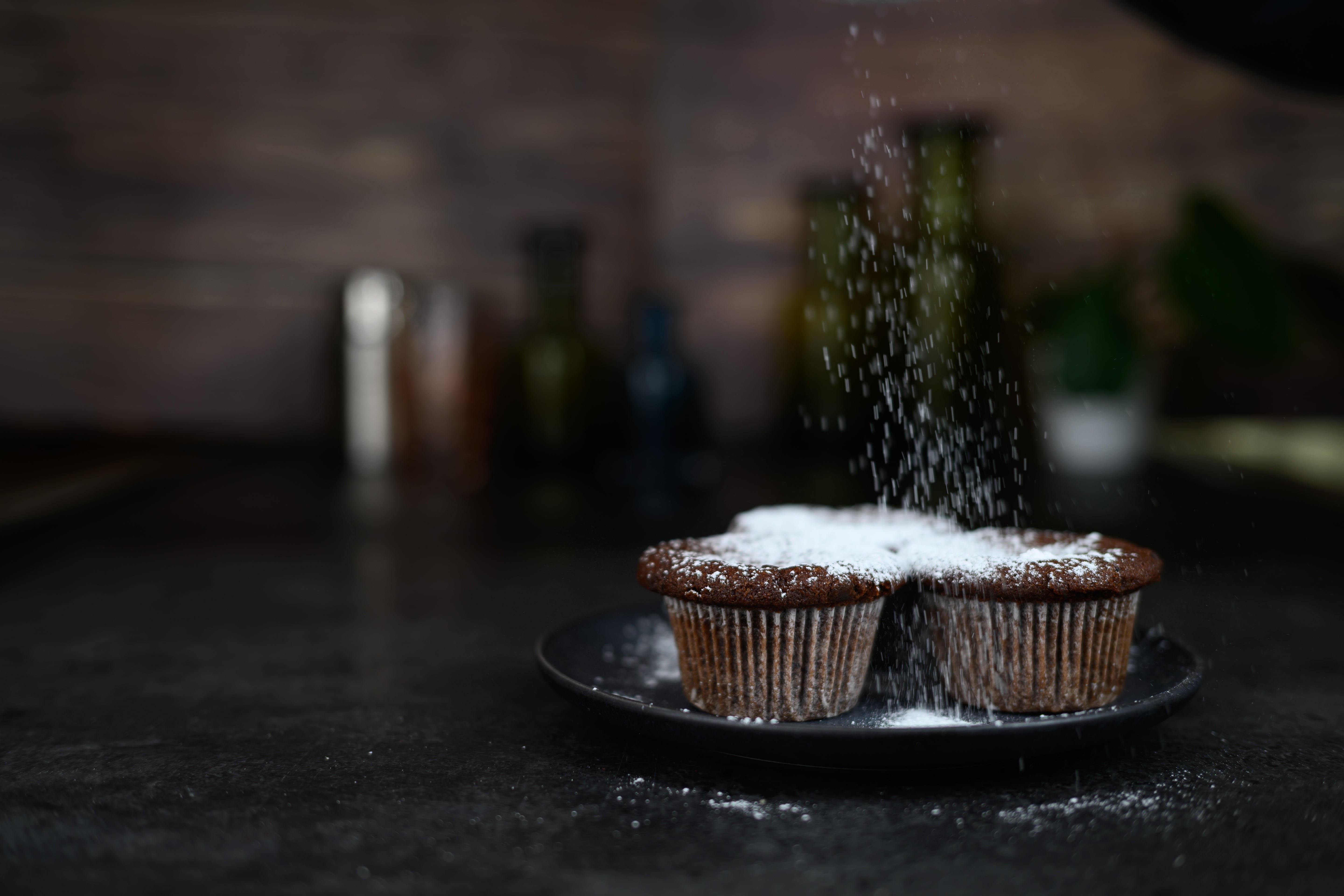 Two cupcakes on a plate with powdered sugar being sprinkled over them in a kitchen setting. Dessert Kitchen Muffin examples